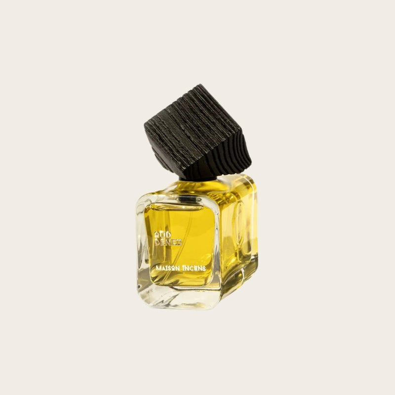 A small Maison Incense perfume bottle with a black ribbed cap, filled with yellow oil, set against a solid off-white background.
